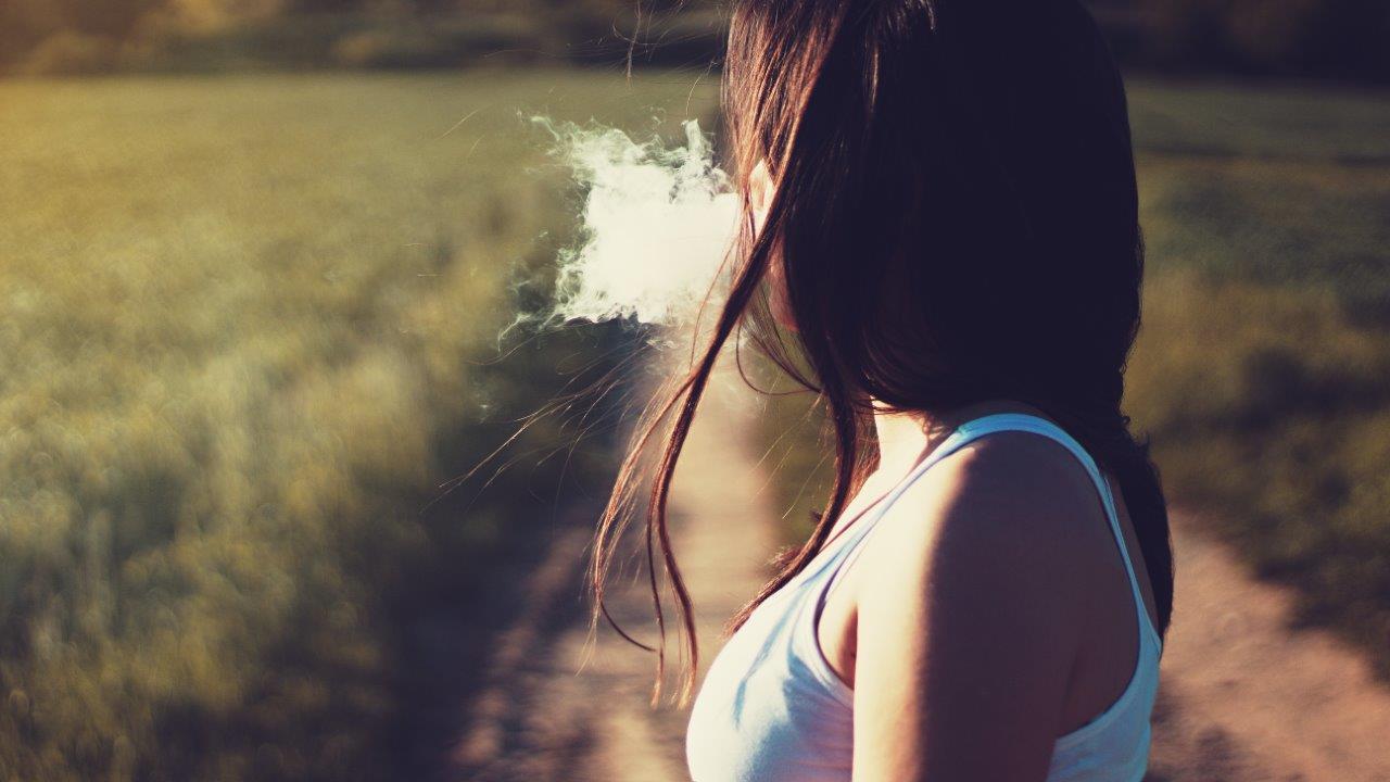 Young person faces away from the camera, as they blow smoke in the middle of a field