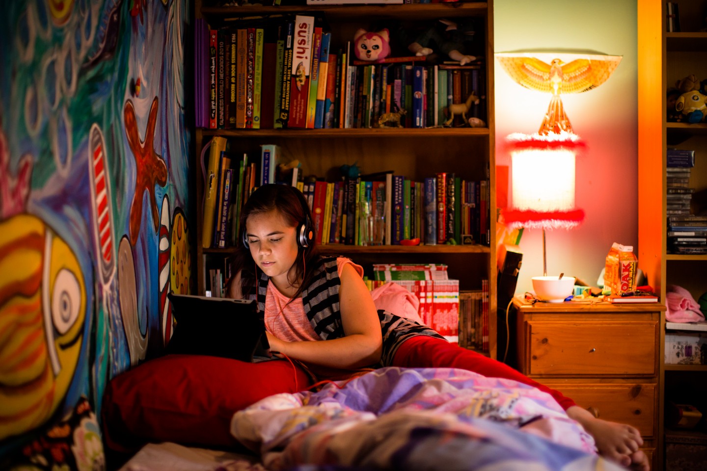 Young person on laptop in front of bookcase in bedroom.